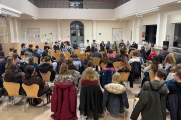 etwa 60 Menschen sitzen in Stuhlreihen und hören einer Gruppe von 14 Personen zu, die ihnen auf Stühlen gegenüber sitzen.